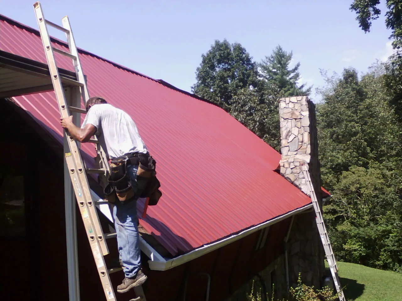 Roofer on a ladder beside a red metal roof and stone chimney — Soffit & Fascia Repair by Roofing Brothers in Declo
